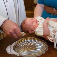 Ein Kind im Taufkleid wird von einer blau gekleideten Frau über eine silberne Schale gehalten, während ein Priester es tauft.